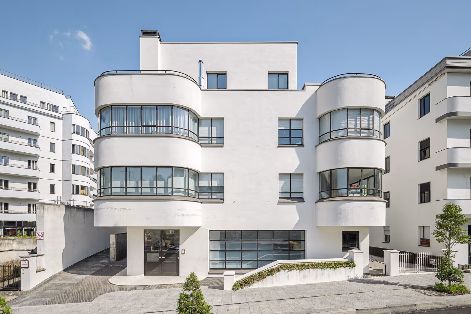 Immeuble moderniste en enduit blanc, quatre niveaux, avec bow-windows courbes en verre et métal noir et balcons arrondis, photographié de face dans un tissu urbain dense sous ciel bleu.
