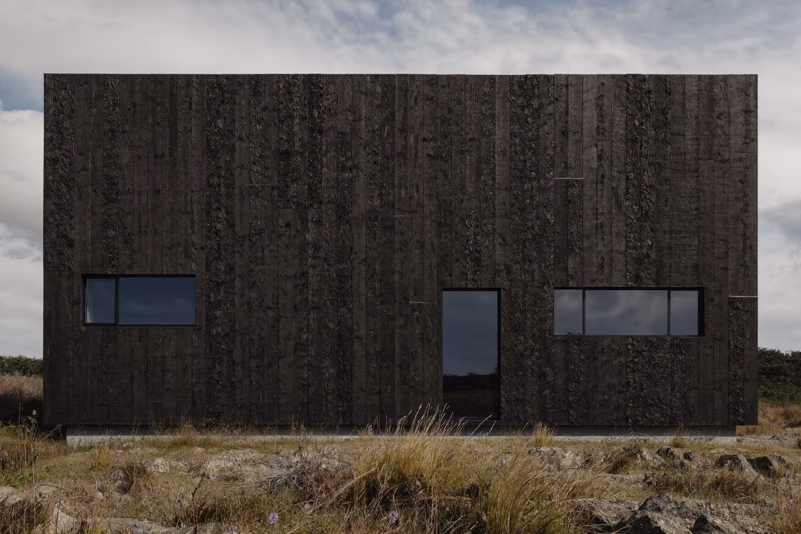 Façade frontale d'un volume cubique recouvert de bardage en bois brûlé noir et texturé, percé de trois ouvertures rectangulaires asymétriques, posé sur un socle en béton dans une lande rocheuse aux herbes sèches.