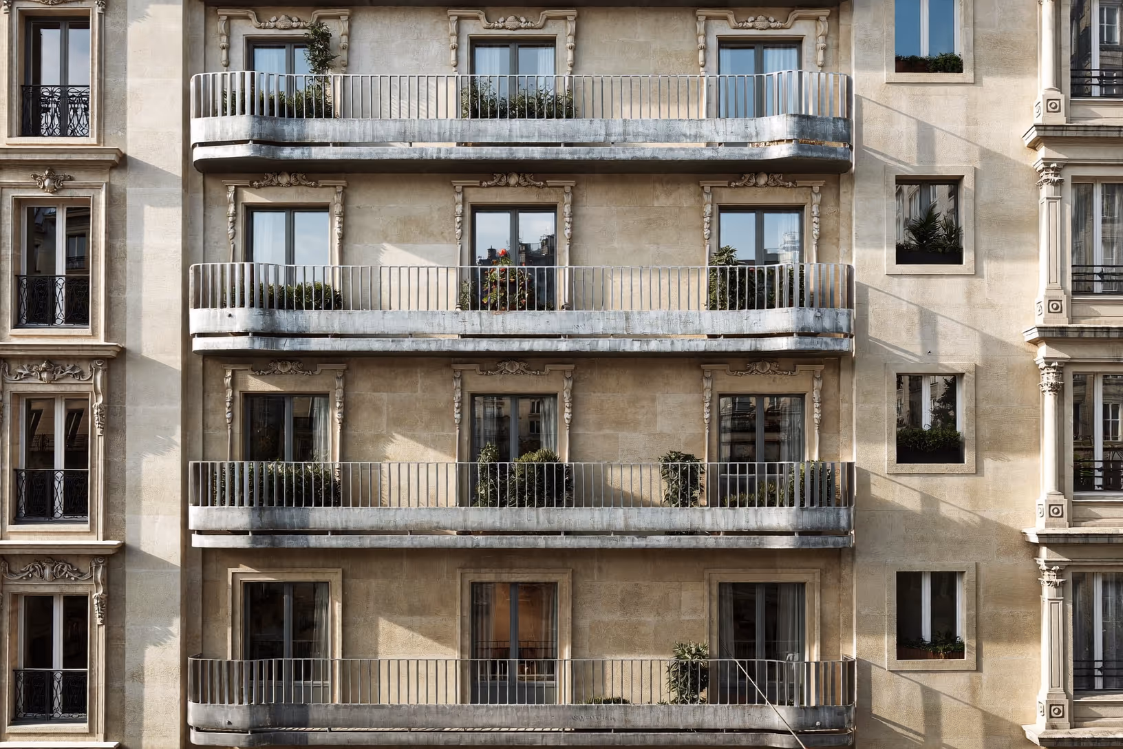 Façade haussmannienne en pierre calcaire beige photographiée frontalement, cinq niveaux de balcons filants aux garde-corps métalliques fins, encadrements sculptés autour des fenêtres à petits carreaux, jardinières vertes, ombres rasantes marquées.