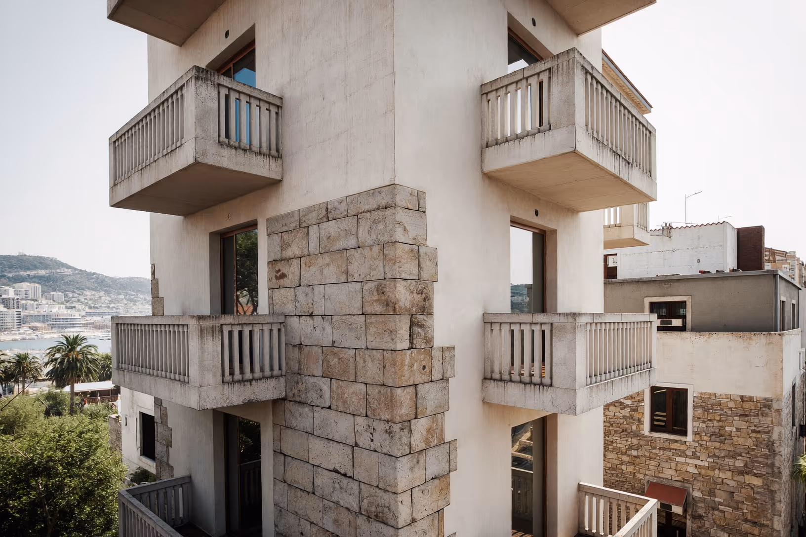 Façade d'un immeuble à plusieurs niveaux composé d'un noyau central en grands blocs de calcaire beige, avec des balcons en béton blanc en porte-à-faux disposés symétriquement de chaque côté, une baie et des collines visibles en arrière-plan.
