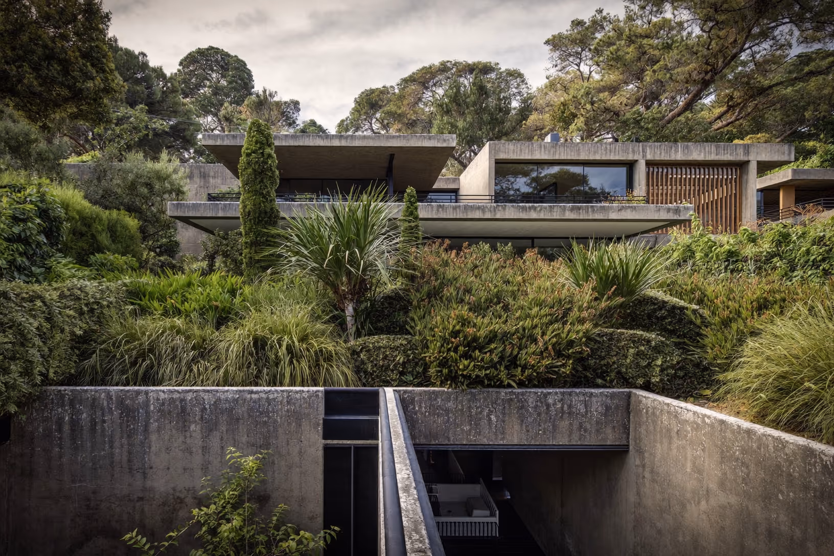 Maison en béton brut étagée sur une pente boisée, avec terrasses horizontales superposées, végétation dense entre les niveaux et couloir encaissé au premier plan.