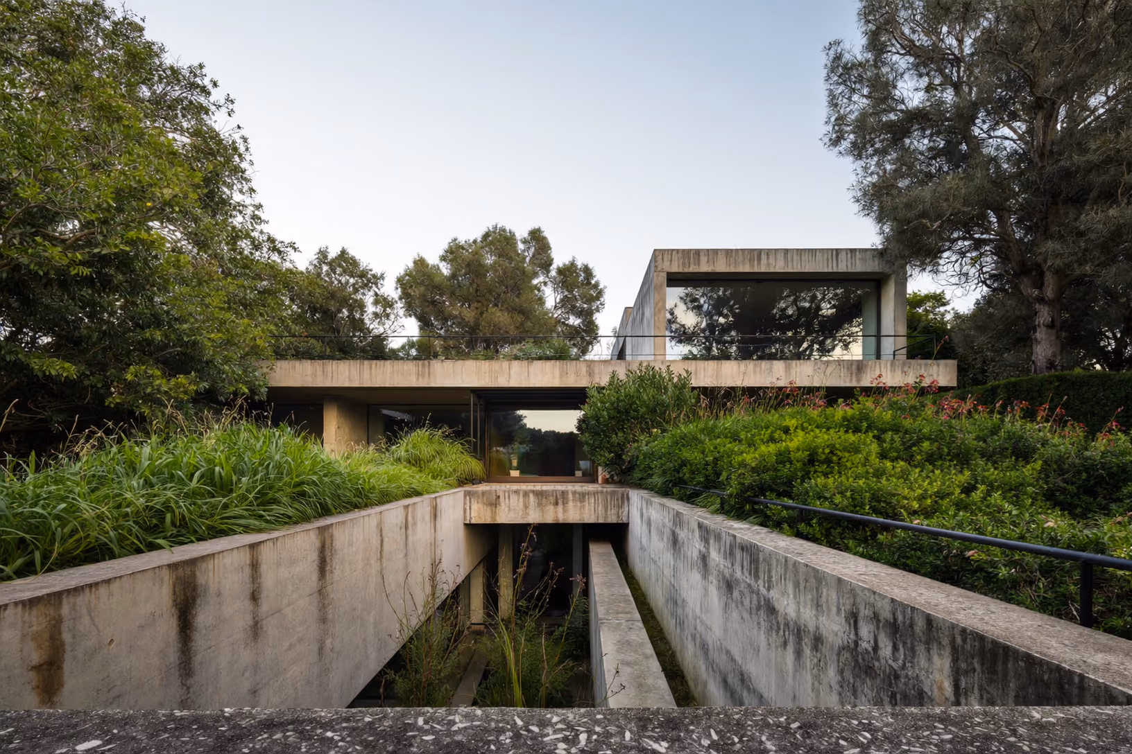Maison en béton brut basse et horizontale enchâssée dans une végétation dense, avec une rampe centrale creusée entre des murs épais et de larges vitrages continus sous des dalles débordantes.