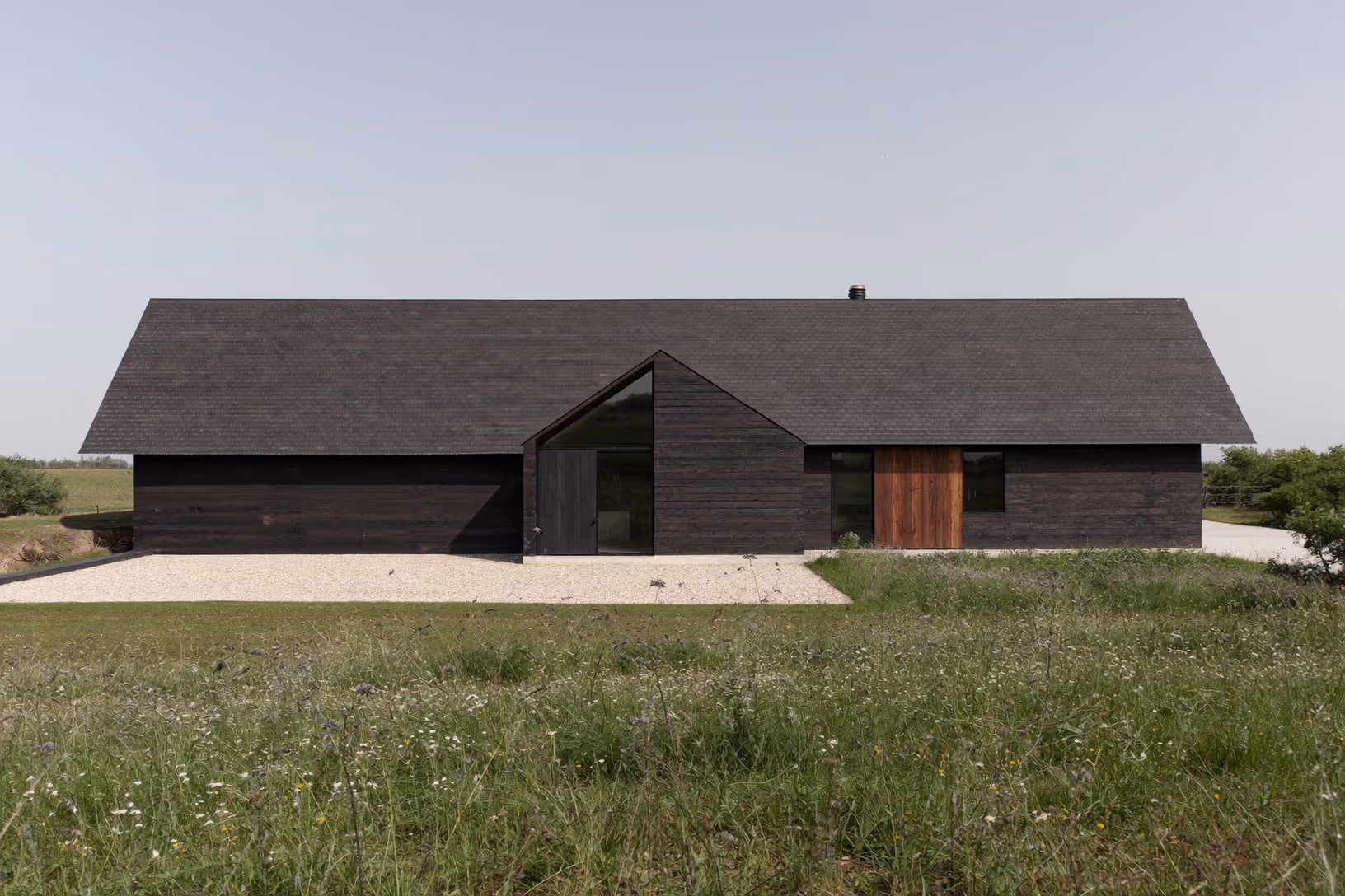 Maison de plain-pied à toiture à deux pans en bardage bois brûlé sombre, porte en bois naturel, grandes ouvertures vitrées en pignon, entourée d'une prairie fleurie sous ciel clair.