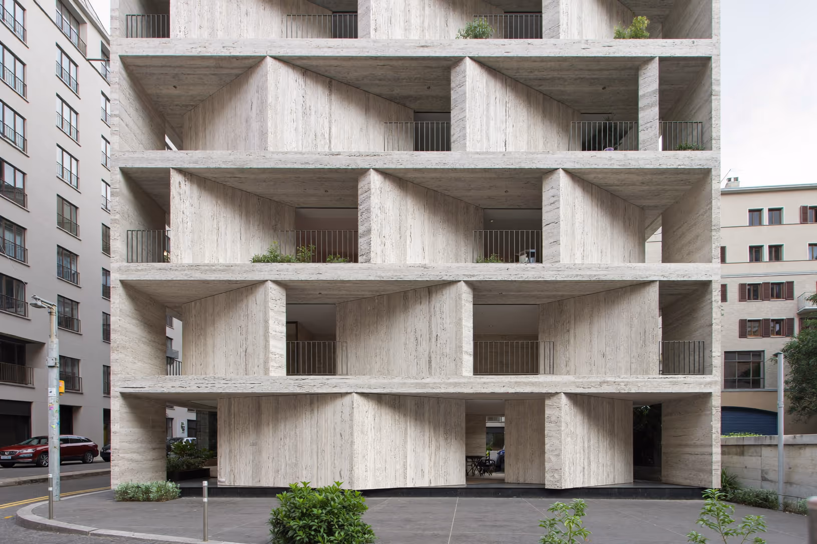 Façade urbaine en travertin clair composée de grandes alvéoles structurelles en saillie, avec balcons filants à garde-corps fins, quelques plantes en terrasse et rez-de-chaussée ouvert sur pilotis.