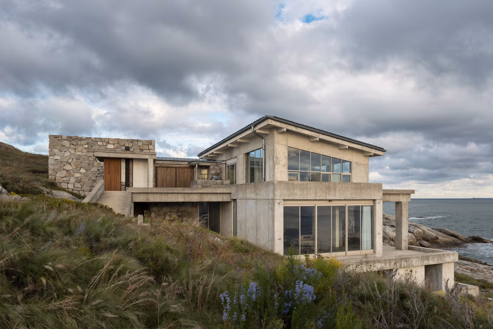 Maison à deux volumes — béton clair et pierre appareillée — implantée en gradins sur un promontoire rocheux côtier, herbes de dune et fleurs mauves au premier plan, mer et ciel nuageux en arrière-plan.