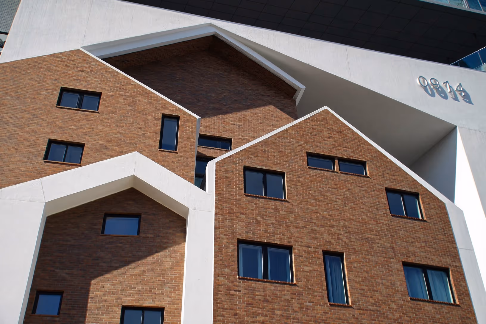Façade d'un immeuble contemporain composée de plusieurs volumes en brique rouge aux toits en pignon, enchâssés dans une structure cadre blanche aux arêtes nettes, photographiée en contre-plongée sous une lumière rasante.