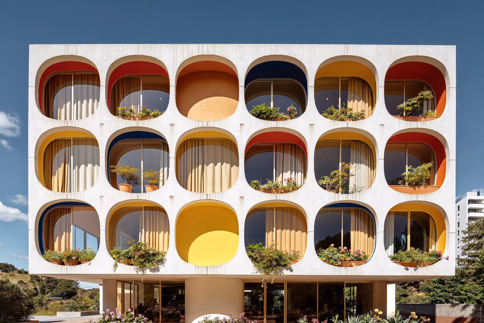 Façade de béton blanc ivoire percée de quinze alvéoles à coins arrondis, disposées en grille régulière sur trois étages, chacune bordée d'un arc de couleur vive — rouge, jaune, bleu marine, orange — avec baies vitrées coulissantes, rideaux ocre et plantes en pot.