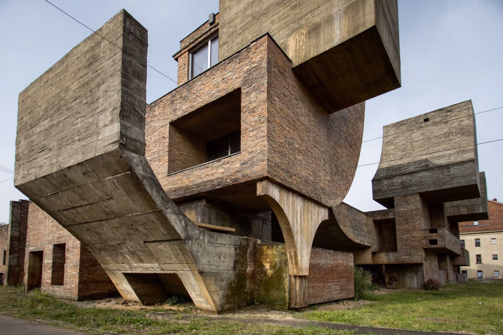 Deux volumes d'habitation brutalistes en béton brut et brique rouge, portés par des piliers courbes massifs, photographiés de face dans un environnement urbain dégradé sous un ciel voilé.