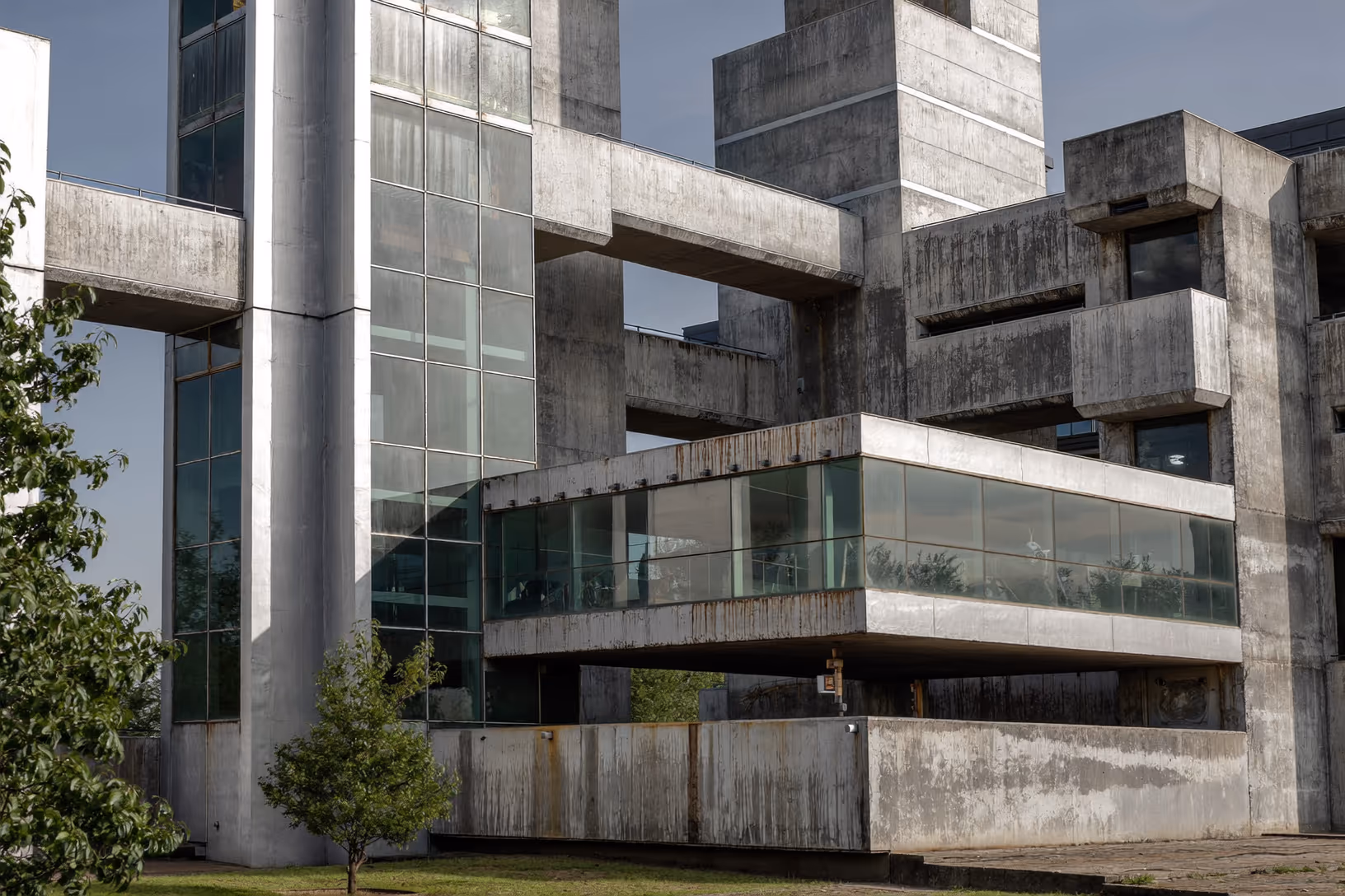 Façade brutaliste en béton brut avec volumes imbriqués, larges bandes vitrées encadrées de poutres épaisses, traces de rouille sur les parois, arbres au premier plan.