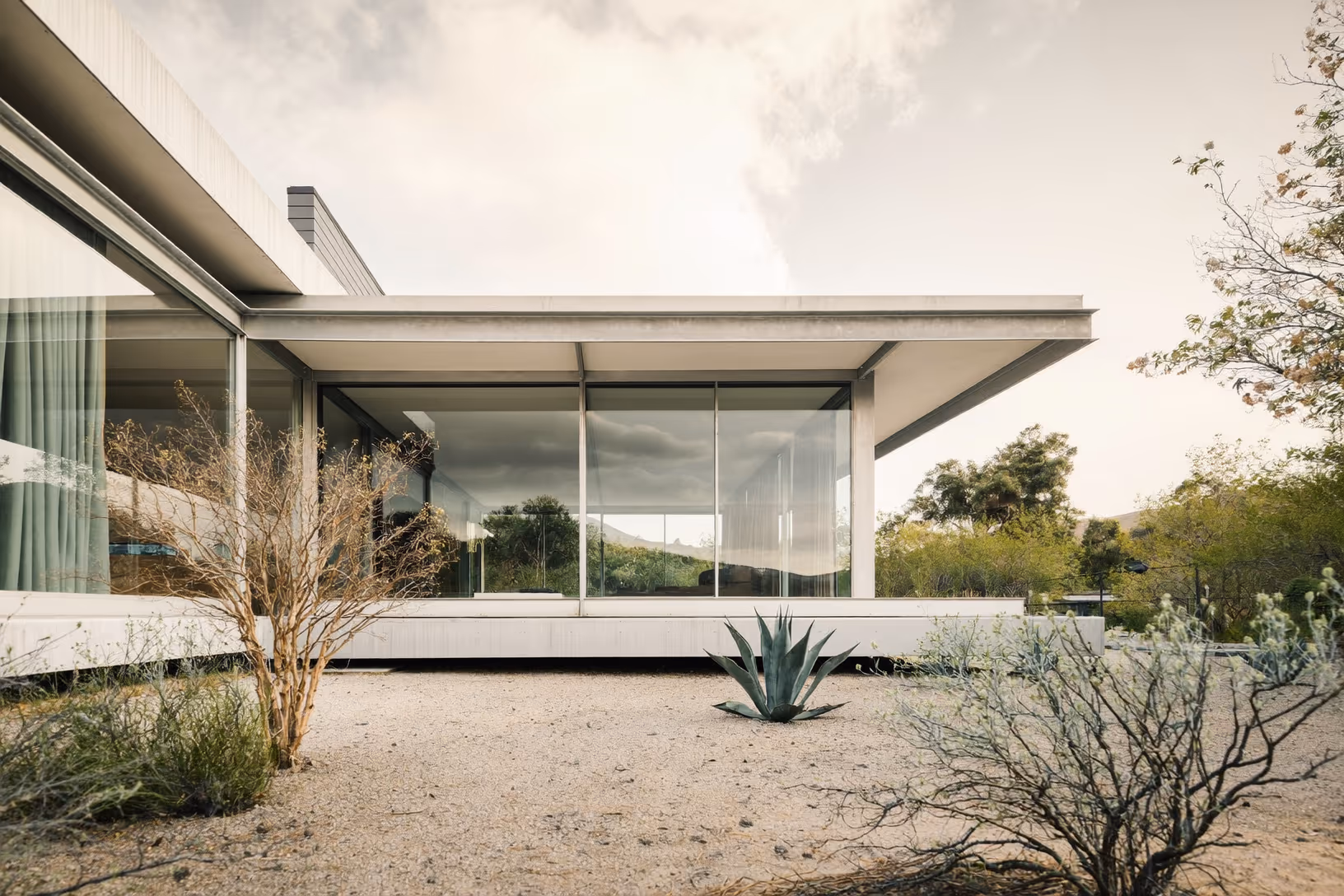 Façade entièrement vitrée d'une maison à toit-dalle horizontal sur socle blanc, entourée d'agaves et d'arbustes désertiques sur fond de ciel couvert et de collines lointaines.