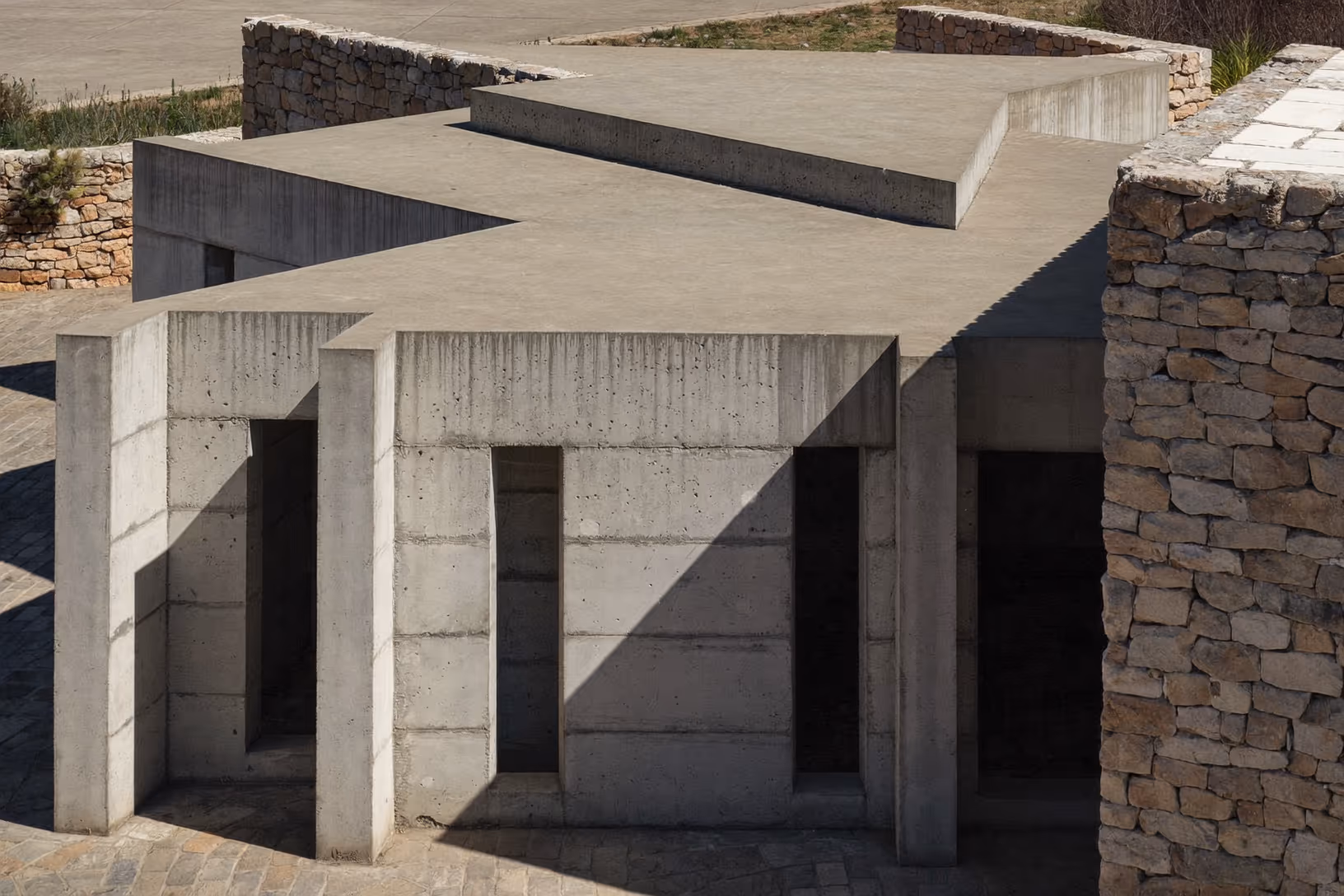 Structure en béton brut avec piliers verticaux et dalle horizontale plate, jouxtant un mur de pierre sèche appareillée, lumière solaire rasante, sol en dalles grises, terrain aride en arrière-plan.