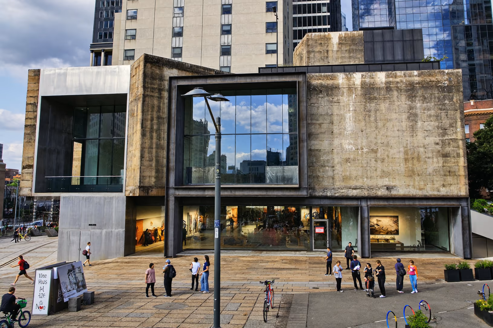 Bâtiment public en béton brut à deux niveaux avec grande baie vitrée centrale encadrée d'un portique métallique sombre, passants sur la place pavée au rez-de-chaussée, tours en verre en arrière-plan urbain.