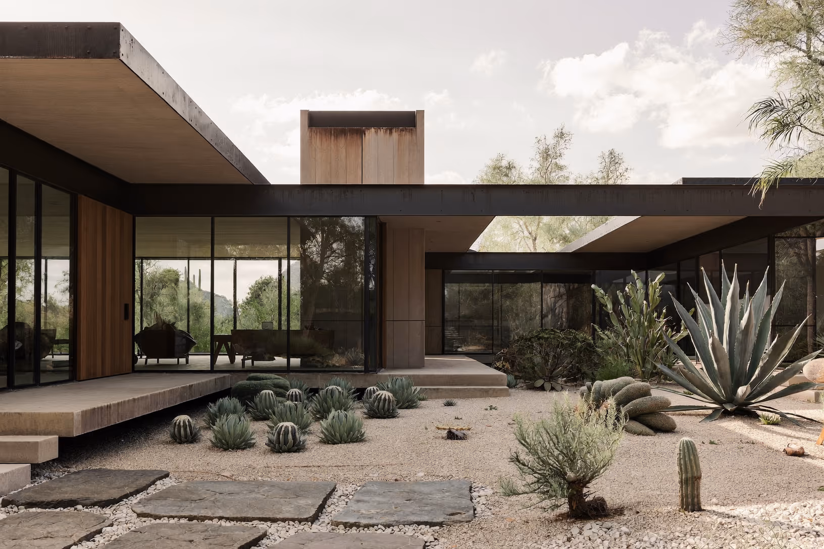Vue extérieure d'une maison de plain-pied à toit plat en acier sombre et verre, avec un volume en bois clair émergeant du toit, entourée d'agaves, de cactus barrel et de saguaros sur un sol de gravier, avec des dalles en pierre brute formant un cheminement.