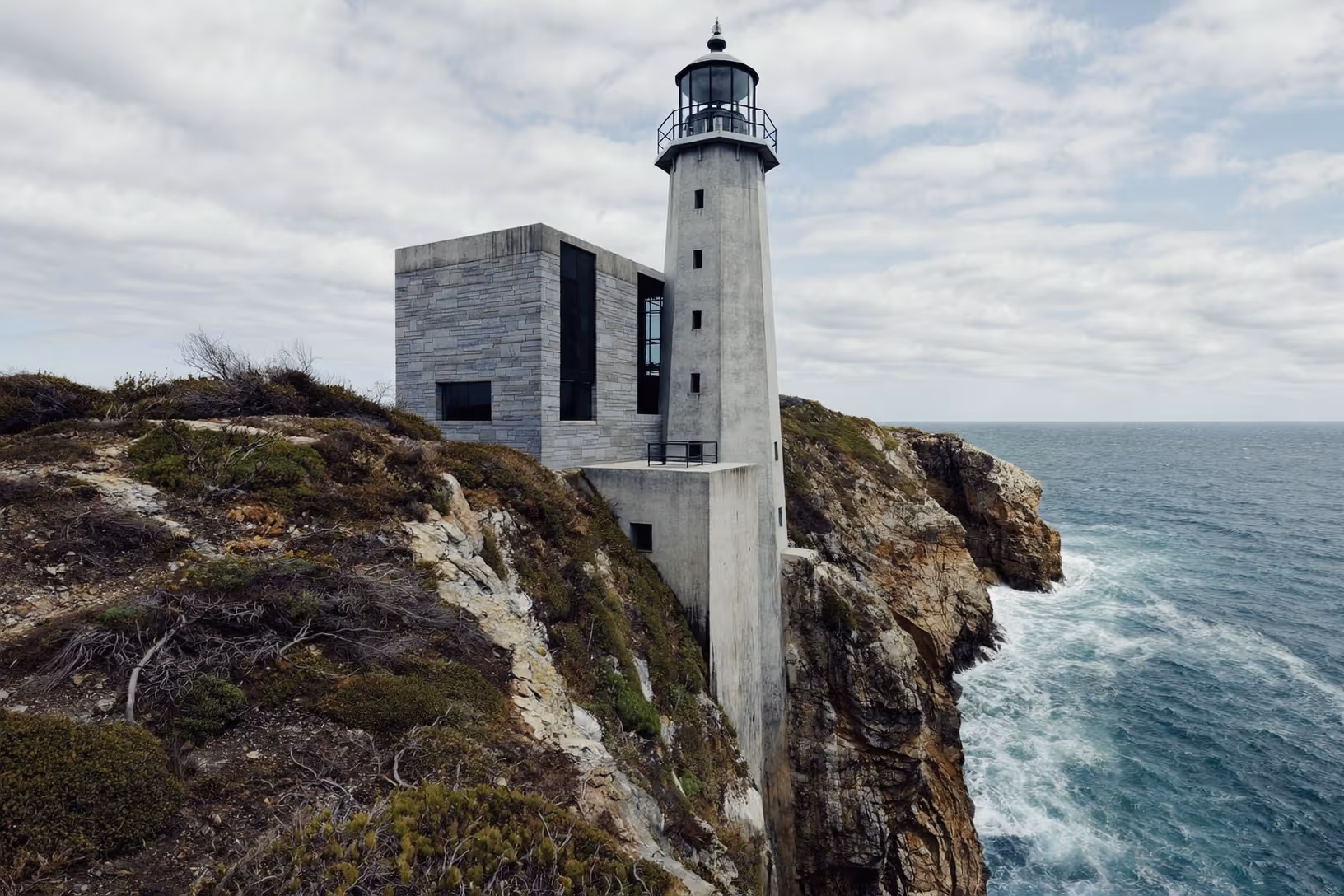 Un phare cylindrique en béton gris et un volume cube en pierre de taille grise, posés au bord d'une falaise rocheuse surplombant une mer agitée, sous un ciel couvert.