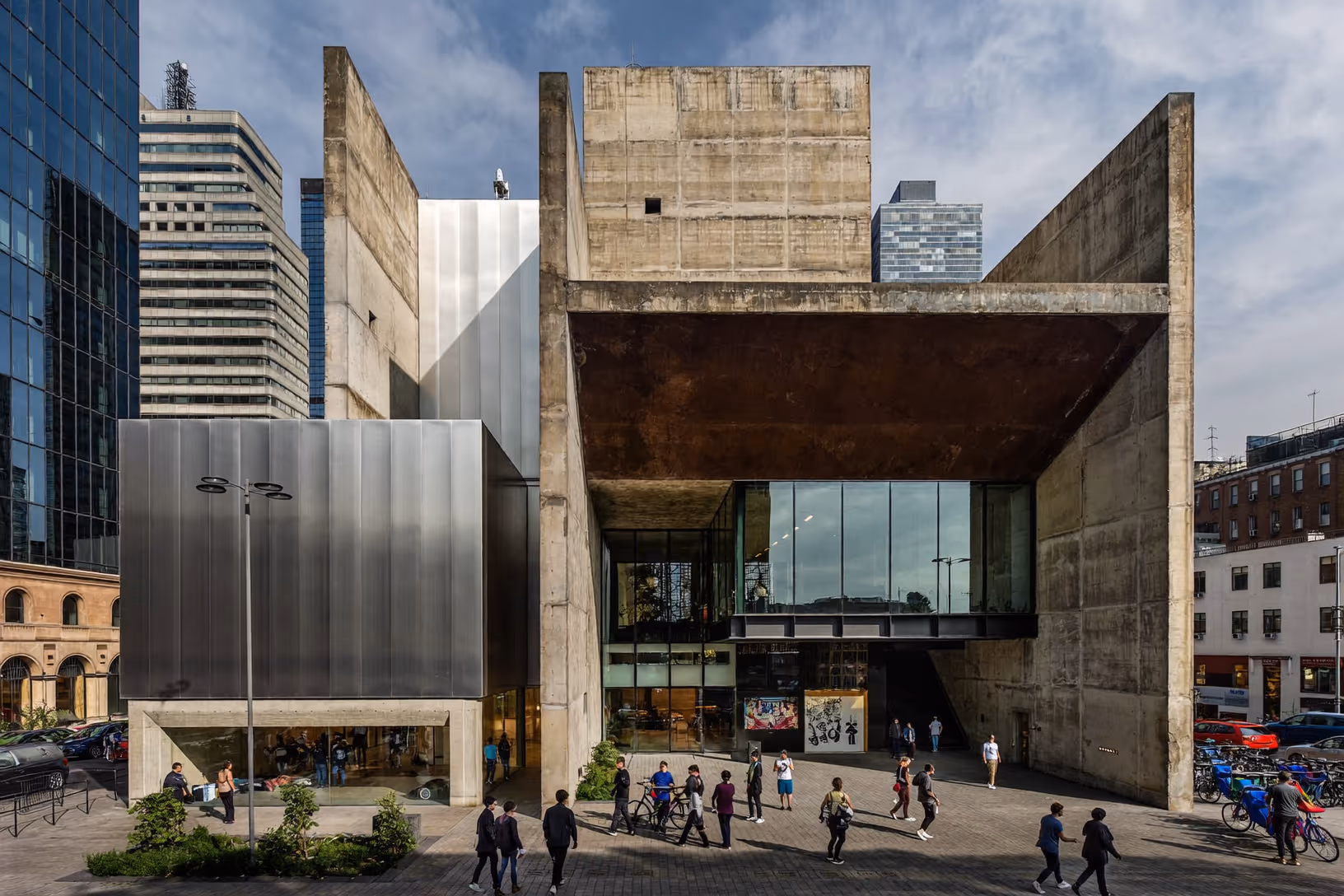 Façade d'un bâtiment brutaliste en béton brut avec une extension en métal brossé argenté à gauche, un porte-à-faux en acier Corten rouille au centre, et une façade vitrée au rez-de-chaussée ; des passants traversent l'esplanade devant le bâtiment.