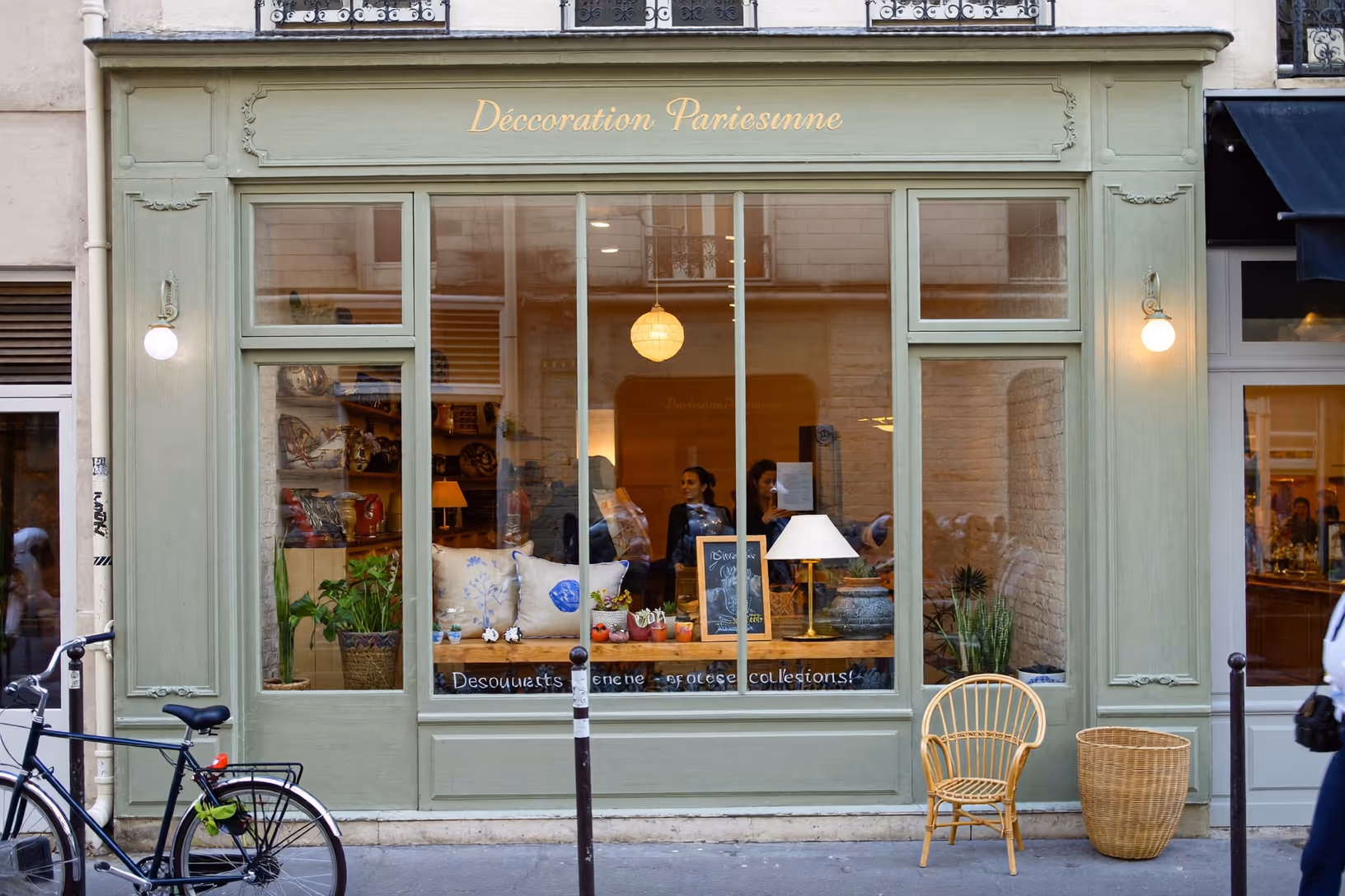 Devanture d'une boutique de décoration parisienne à la façade vert sauge, vitrine éclairée garnie d'objets de déco, un vélo et un fauteuil en rotin sur le trottoir.