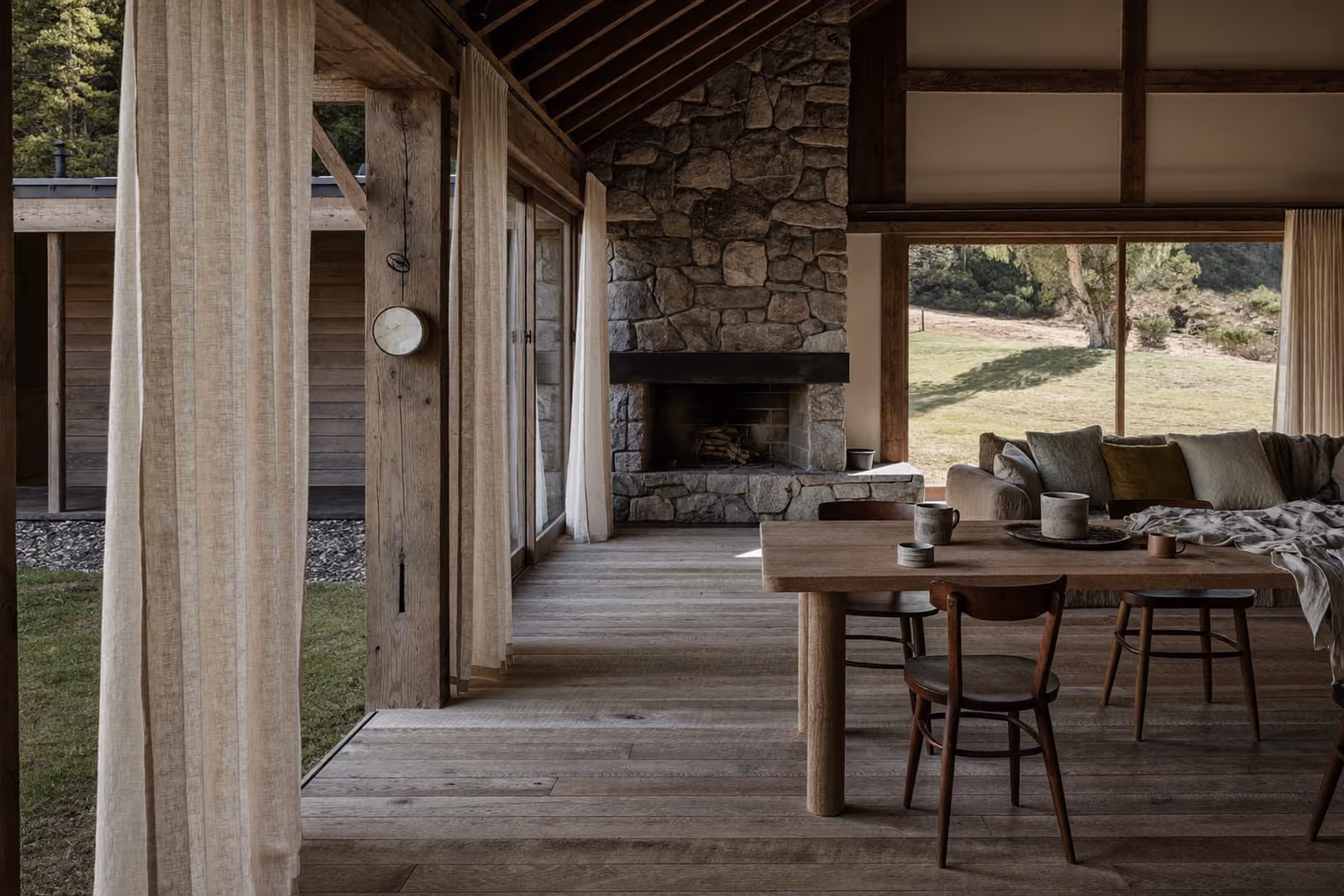 Véranda Japandi aux rideaux de lin, cheminée de pierre et table en bois ouverte sur le jardin.