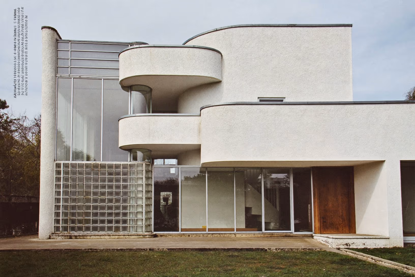 Villa moderniste blanche d'inspiration Bauhaus : volumes cubiques et balcons arrondis à garde-corps de verre courbes, mur en briques de verre, larges baies vitrées et porte en bois, toiture plate, sur une pelouse.